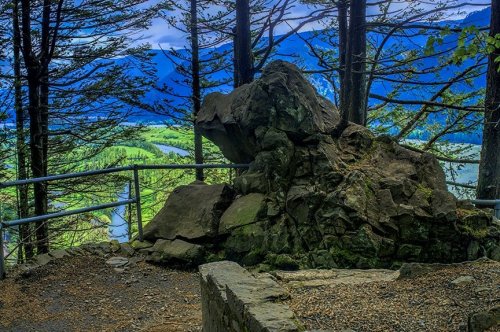 A rocky outcrop overlooks a lush green valley and winding river, framed by tall trees under a cloudy sky.