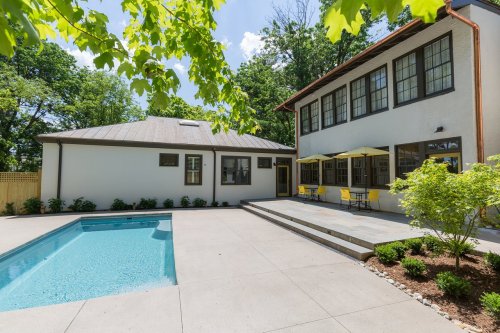 A modern backyard with a swimming pool, patio, and a two-story house surrounded by greenery.