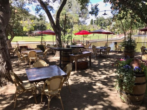 Outdoor dining area with wooden tables, chairs, and umbrellas, surrounded by trees and a scenic view of a pond.