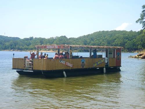 A tour boat with a thatched roof is on a lake, surrounded by green hills and people enjoying the ride.