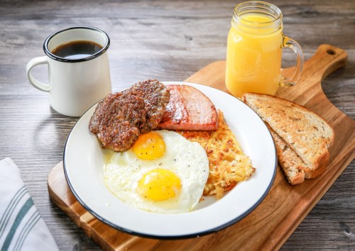 A hearty breakfast plate with eggs, sausage, ham, hash browns, toast, coffee, and orange juice on a wooden board.