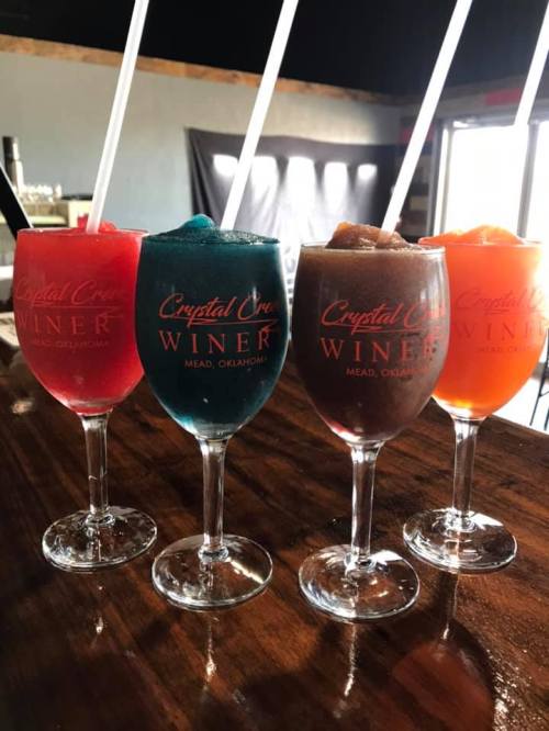 Four colorful frozen drinks in glasses with "Crystal Creek Winery" logo, sitting on a wooden bar.