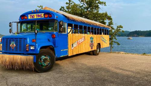 A blue bus with a tiki theme, featuring a thatched roof and wooden decor, parked by a lake.