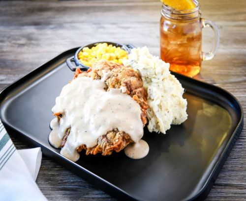 A plate of fried chicken smothered in gravy, served with mashed potatoes, corn, and a glass of iced tea.