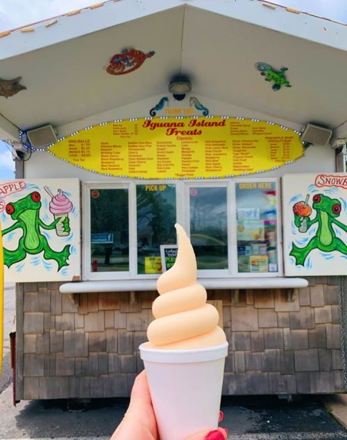 A hand holds a soft-serve ice cream cone in front of a colorful ice cream stand with a menu.