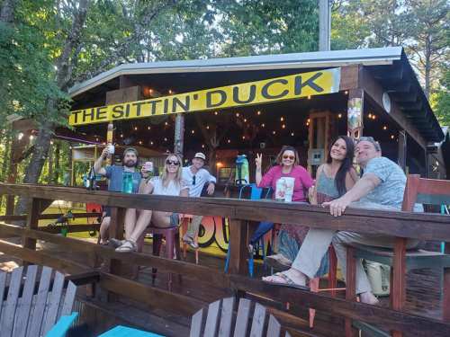A group of six people enjoying drinks on a deck in front of a colorful bar named "The Sittin' Duck" surrounded by trees.