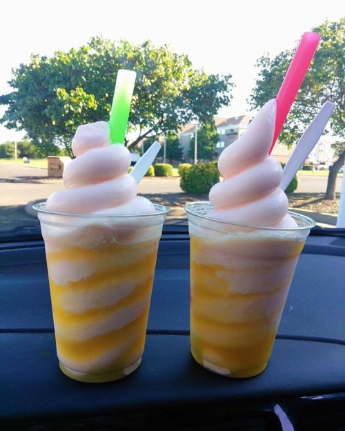 Two colorful frozen drinks with swirled soft serve, served in clear cups with green and pink straws, on a car dashboard.