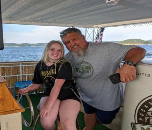 A man and a girl smile together on a boat, with water and hills in the background. The girl is sitting on a stool.