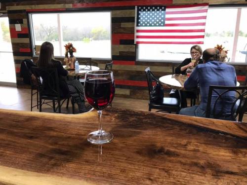 A glass of red wine on a wooden table, with people seated at tables and an American flag in the background.