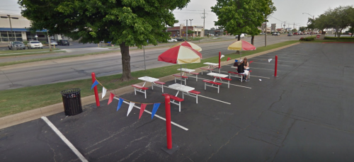 Outdoor dining area with red and yellow umbrellas, tables, and decorative flags beside a parking lot.