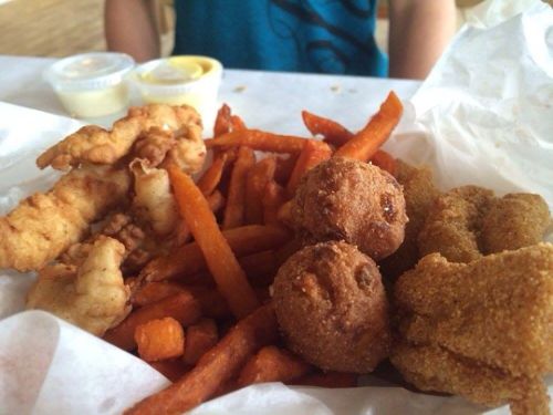 A plate of fried chicken strips, sweet potato fries, and hushpuppies, with dipping sauces in the background.