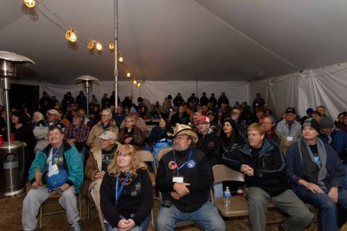 A large audience seated under a tent, listening attentively, with warm lighting and heaters in the background.