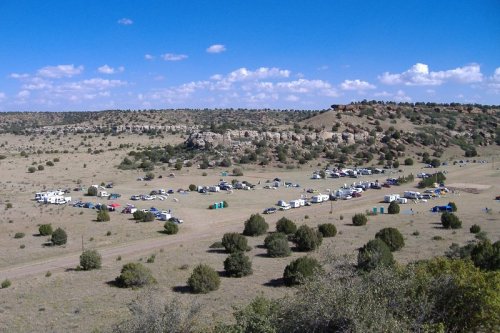 A wide view of a campsite in a desert landscape, with tents and vehicles scattered across a grassy area under a blue sky.