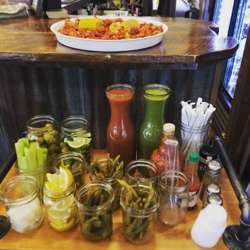 A wooden table displays a platter of crawfish and jars of garnishes, including celery, lemons, and sauces.