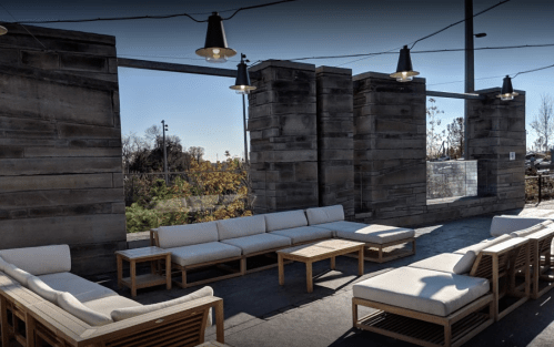 Outdoor seating area with modern furniture, stone walls, and string lights, surrounded by greenery.