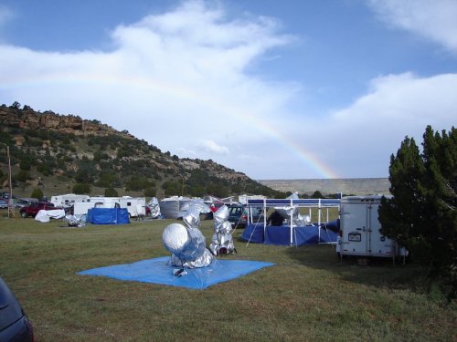 A campsite with silver tarps and trailers under a rainbow and blue sky, surrounded by hills and greenery.