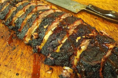 Sliced, smoky barbecue meat with a dark crust and barbecue sauce, resting on a wooden cutting board next to a knife.