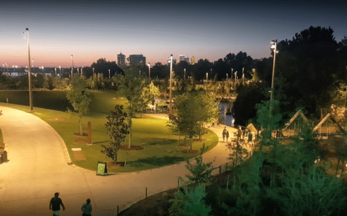 A park at dusk with walking paths, trees, and city skyline in the background, illuminated by soft lights.