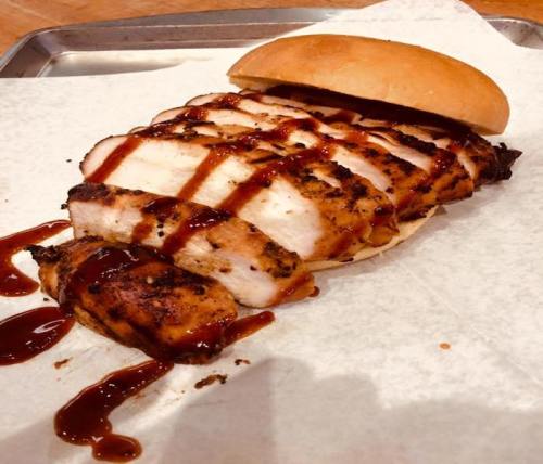 Grilled chicken breast sliced and drizzled with barbecue sauce, served with a bun on parchment paper.