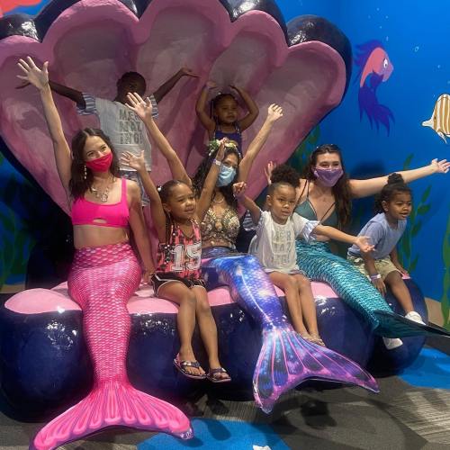 A group of children and adults in mermaid costumes pose joyfully in a large seashell backdrop.