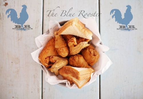 A basket of crispy fried chicken pieces on a wooden table, with "The Blue Rooster" logo in the background.