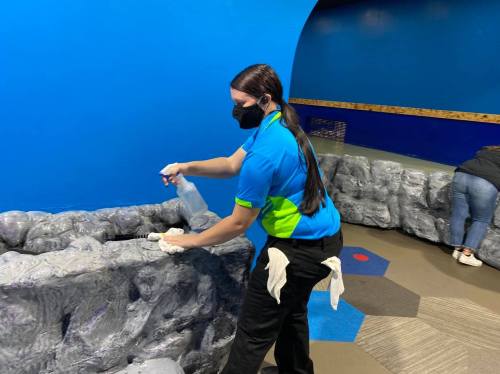A staff member in a blue and green uniform sprays a surface while wearing a mask in a colorful indoor space.