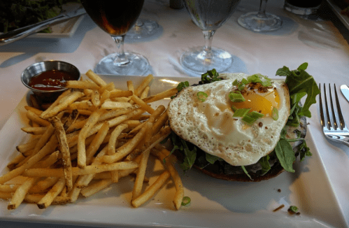 A plate with a burger topped with a fried egg, greens, and a side of crispy fries and ketchup.