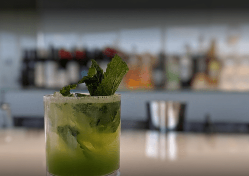 A refreshing green cocktail with mint leaves, served in a glass on a bar counter with blurred bottles in the background.