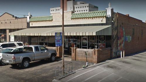 A brick building with a green awning, featuring outdoor seating and parked cars in front.
