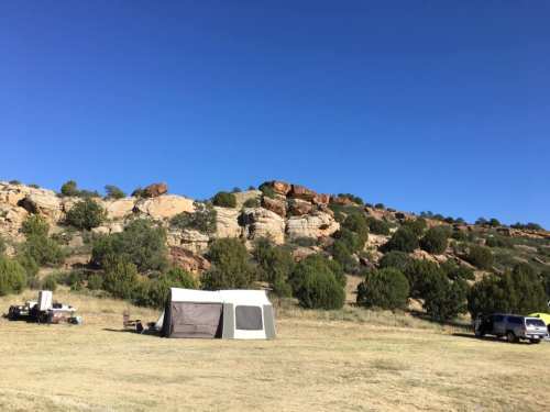 A campsite with a tent set up on grassy land, surrounded by rocky hills and sparse trees under a clear blue sky.