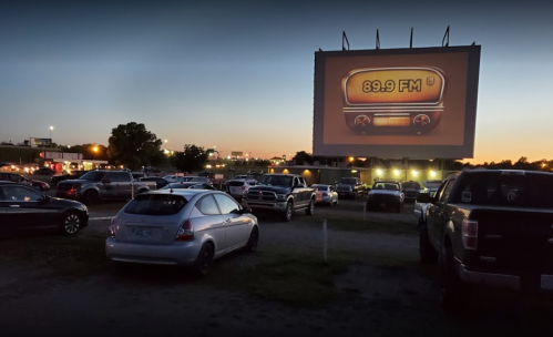 A drive-in theater scene at dusk, featuring a large screen displaying "89.9 FM" and parked cars in the foreground.