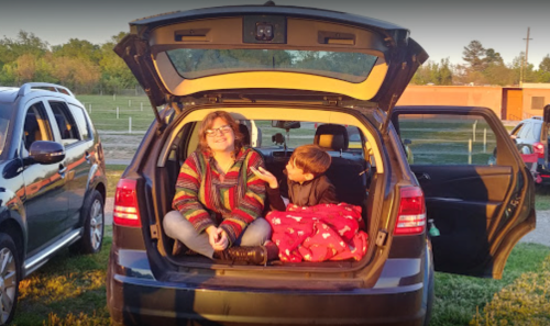 A woman and a child sit in the back of a car, enjoying the sunset, surrounded by parked vehicles.