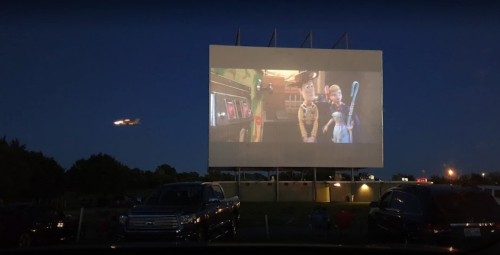 A drive-in movie screen shows animated characters at night, with cars parked in front and an airplane flying above.