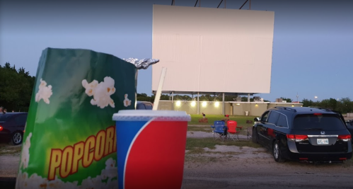 A close-up of popcorn and a drink in front of a drive-in movie screen at dusk, with cars and chairs in the background.
