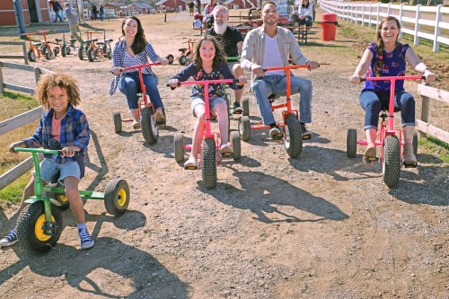 A group of six people riding colorful tricycles on a dirt path at a farm, smiling and enjoying a sunny day.