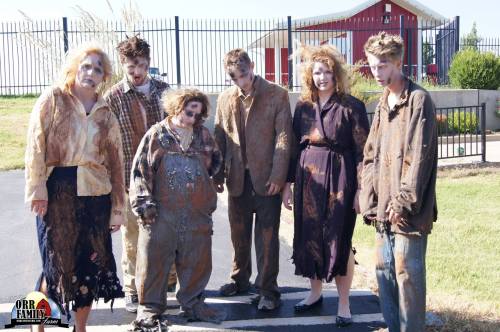 A group of six people dressed as zombies, covered in dirt and tattered clothing, posing outdoors near a fence.