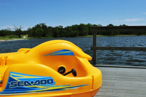 A bright yellow Sea-Doo watercraft on a dock by a calm lake, surrounded by trees and a clear blue sky.