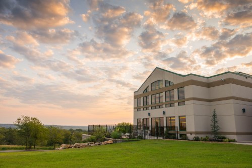 A modern building with large windows, set against a colorful sunset and cloudy sky, surrounded by green grass and trees.