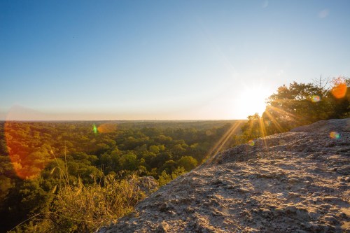 A sun setting over a lush green landscape, viewed from a rocky outcrop, with lens flare effects.