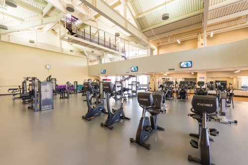 A spacious gym with various exercise machines and equipment, bright lighting, and large windows.