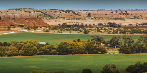 A scenic view of rolling green fields and distant red cliffs under a clear blue sky.