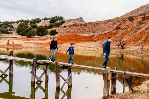 Three children walking on a wooden bridge over a calm lake, surrounded by grassy hills and red earth.