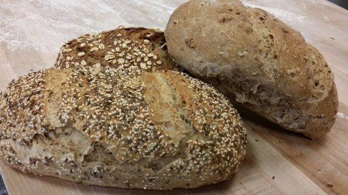 Two loaves of freshly baked bread, one topped with oats and seeds, resting on a wooden surface.