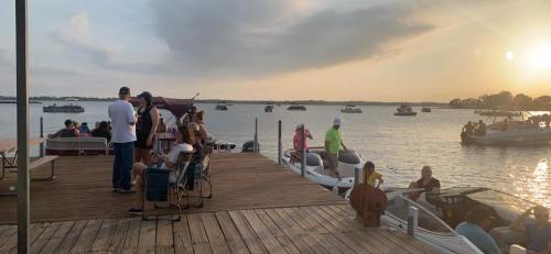 A lively lakeside scene at sunset with people on a dock and boats on the water.