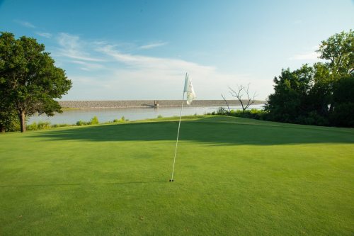 A serene golf course with a flag on the green, overlooking a calm body of water and a distant dam under a blue sky.