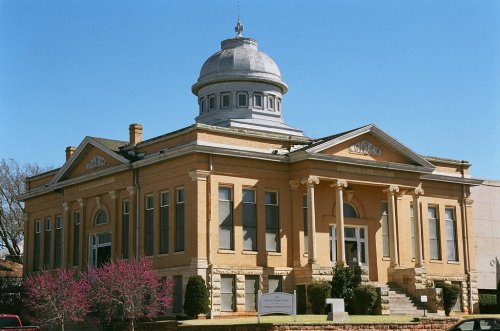 Historic building with a dome, large windows, and columns, surrounded by greenery and a clear blue sky.