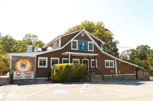A brown building with a sign, surrounded by trees and a parking lot, featuring a logo and accessible parking spaces.