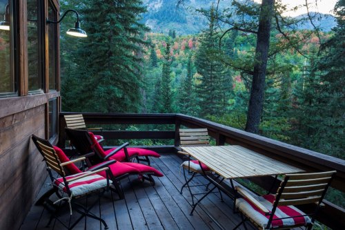 A cozy wooden deck with red lounge chairs and a table, surrounded by lush green trees and mountains in the background.