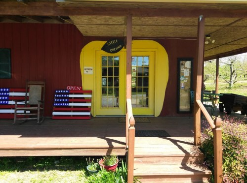 A red building with a yellow door and an "Open" sign, featuring American flag chairs on the porch.
