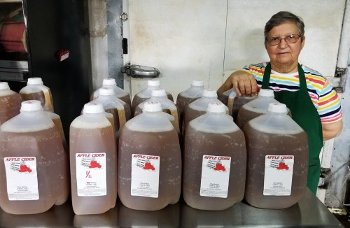 A woman in an apron stands beside several jugs of apple cider on a table in a food preparation area.